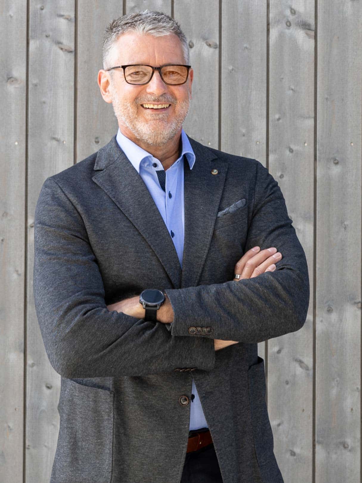 Porträt von Dr. Markus Blaschka vor einer Holzfassade – lächelnd, im Sakko mit verschränkten Armen